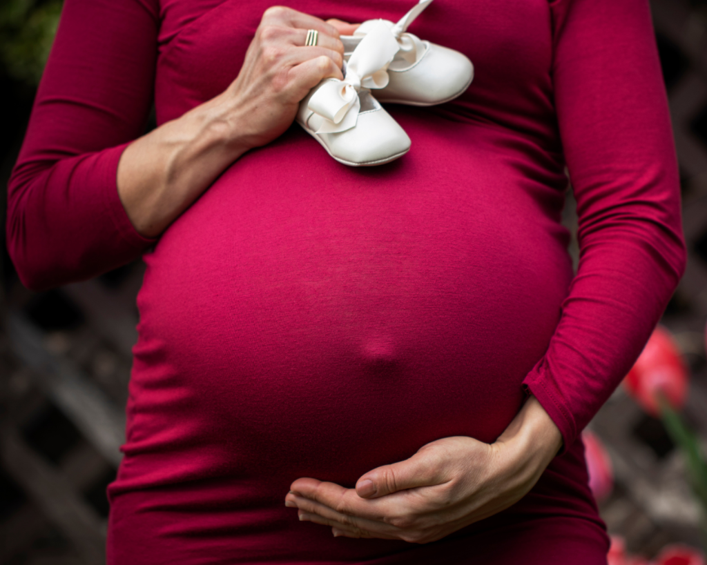 photo showing a pregnant woman in a red dress holding her belly and a pair of baby shoes