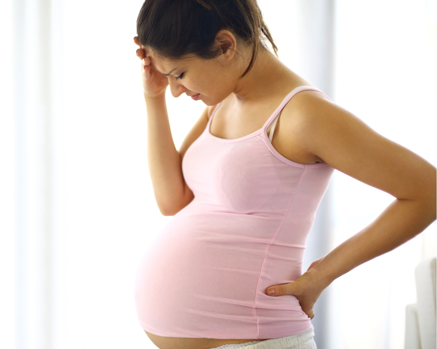 Pregnant woman in a pink tank top with one hand holding her back and her other hand holding her head